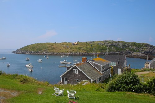 A scenic coastal view featuring boats in the water, grassy hills, and a charming house with white chairs in the foreground.
