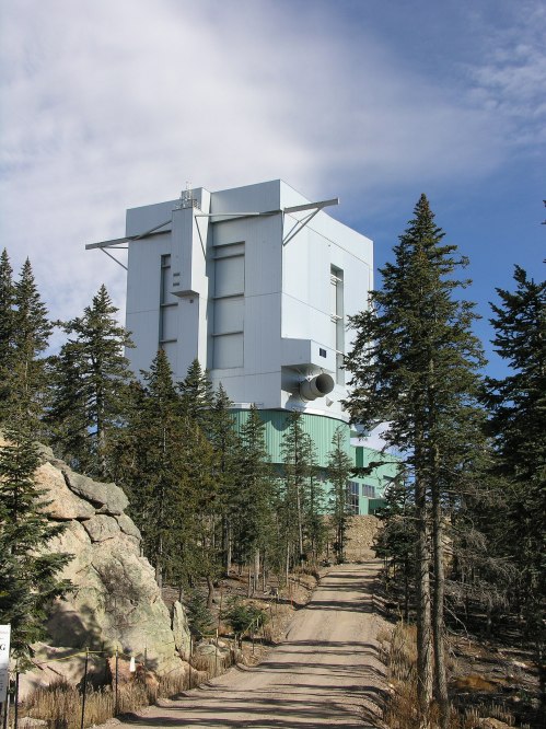 A tall, modern building surrounded by trees on a dirt road, set against a blue sky with scattered clouds.