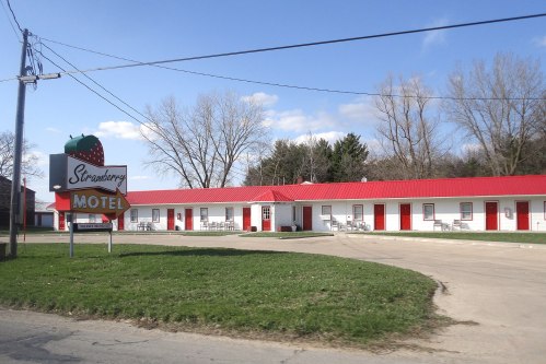 A roadside motel with a red roof and strawberry-themed sign, surrounded by green grass and trees under a blue sky.