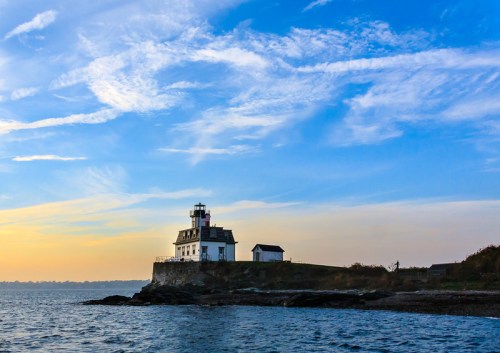 A lighthouse stands on a rocky shore under a blue sky with wispy clouds, reflecting the warm glow of sunset.