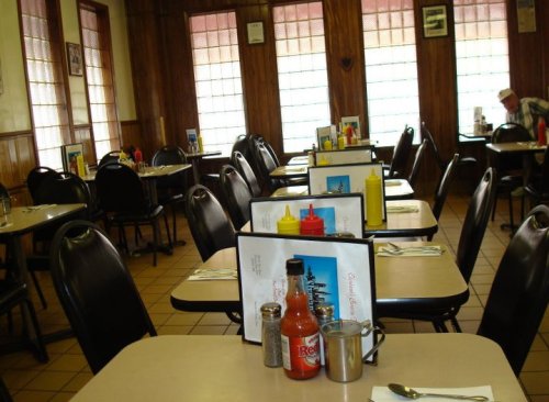 A cozy diner interior with empty tables, condiments, and menus, featuring wooden paneling and large windows.