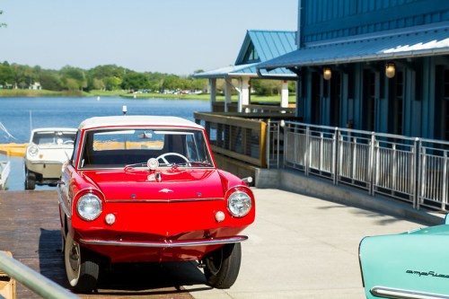 A bright red vintage car parked by a lake, with a blue building and another boat in the background.
