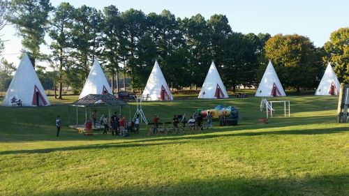A grassy area with several teepee-shaped structures, playground equipment, and people enjoying the outdoors.