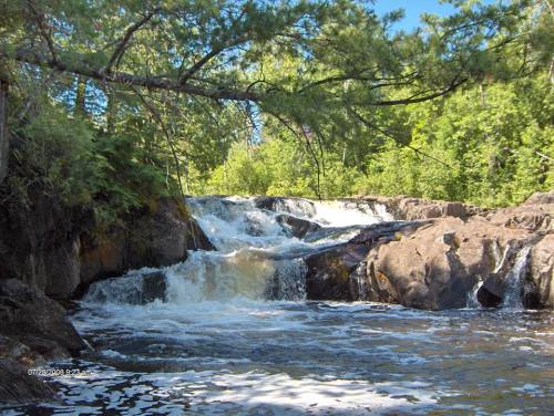 A serene waterfall cascades over rocky terrain, surrounded by lush green trees under a clear blue sky.