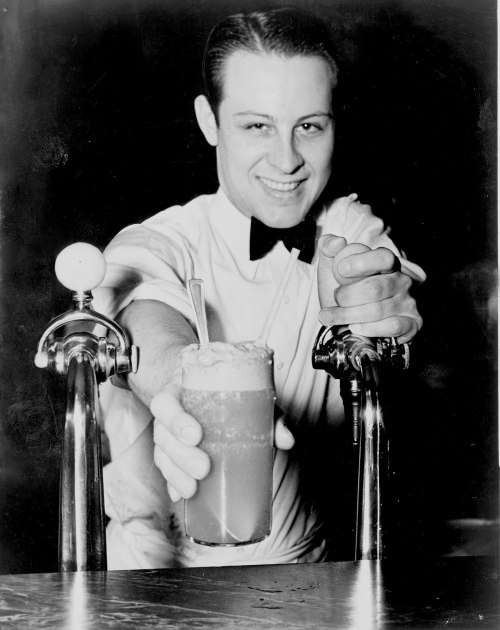A smiling bartender in a bow tie serves a frothy drink from a soda fountain.