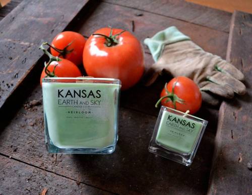 A green candle labeled "Kansas Earth and Sky" beside fresh tomatoes and a gardening glove on a wooden surface.