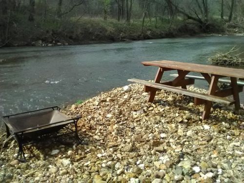 A picnic table by a riverbank, with a fire pit nearby, surrounded by pebbles and trees in the background.