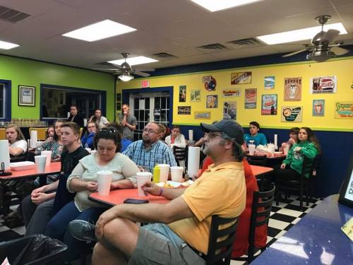 A group of people sitting at tables in a colorful diner, watching a presentation or event.