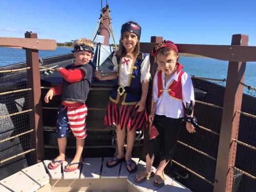 Three children dressed as pirates stand on a boat, smiling against a clear blue sky and calm water.