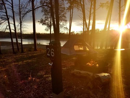 A campsite by a lake at sunset, featuring a tent, campfire, and a sign with the number 26.