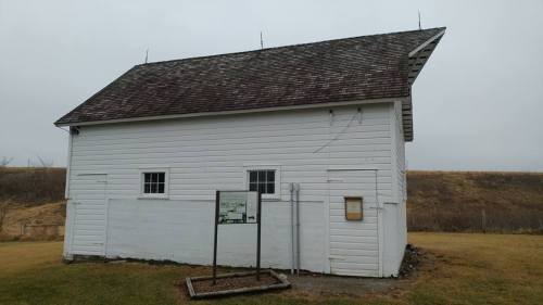 A white wooden building with a sloped roof, featuring small windows and a sign in front, set in a grassy area.