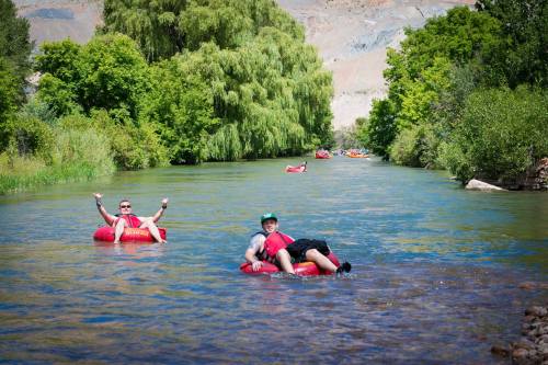 Two people float on inner tubes in a river, surrounded by lush greenery and other tubers in the background.