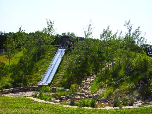 A metal slide on a grassy hill surrounded by trees and a rocky path, under a clear blue sky.