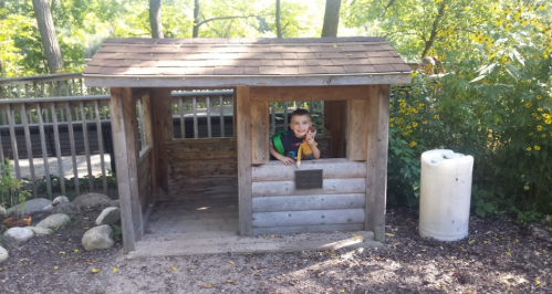 A child peeks out from a wooden playhouse surrounded by trees and flowers in a sunny outdoor setting.