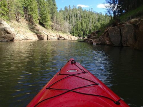 A red kayak on calm water, surrounded by lush green trees and rocky cliffs under a clear blue sky.