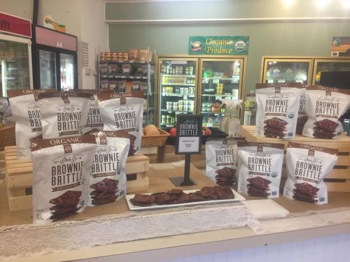 Display of organic brownie brittle bags on a wooden table in a store, with a sign highlighting the product.