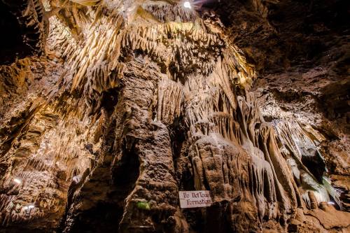 Stalactites and stalagmites in a dimly lit cave, showcasing intricate rock formations. A sign reads "Do Not Touch."