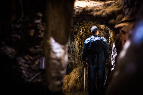A person with a backpack stands in a narrow, dimly lit cave, facing away from the camera.