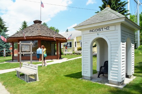 A woman stands near a gazebo with flags, beside a historical marker and a small white structure in a grassy area.