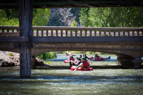People rafting under a bridge on a river, surrounded by greenery and rocky banks on a sunny day.