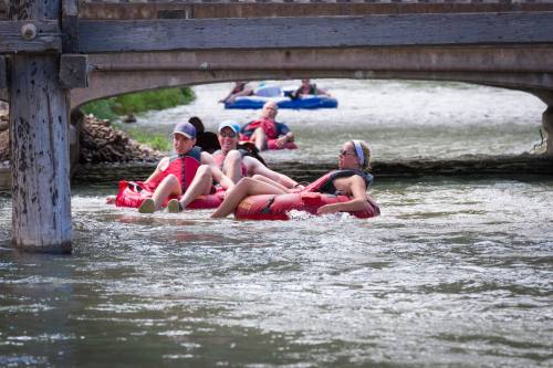 People floating on inner tubes in a river, passing under a wooden bridge on a sunny day.