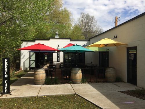 Outdoor seating area of Fresco Cafe with colorful umbrellas, surrounded by greenery and a light-colored building.