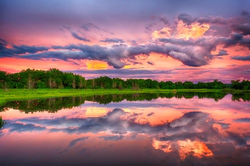 A serene lake at sunset, reflecting vibrant clouds and colorful skies, surrounded by lush greenery.