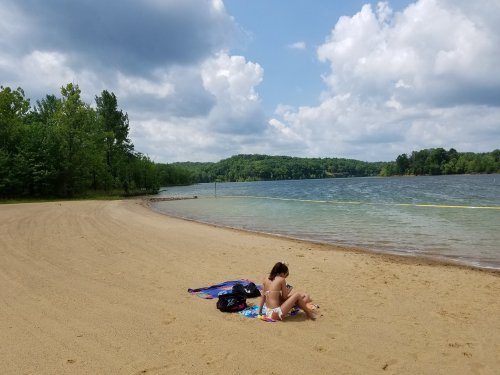A person sits on a beach towel by a calm lake, surrounded by trees and under a partly cloudy sky.