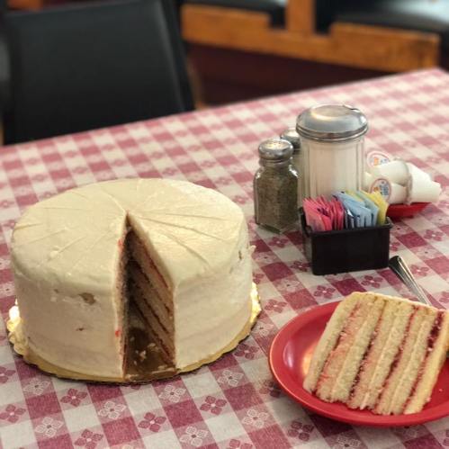 A round cake with white frosting and a slice cut out, placed on a checkered tablecloth next to a plate of cake slices.