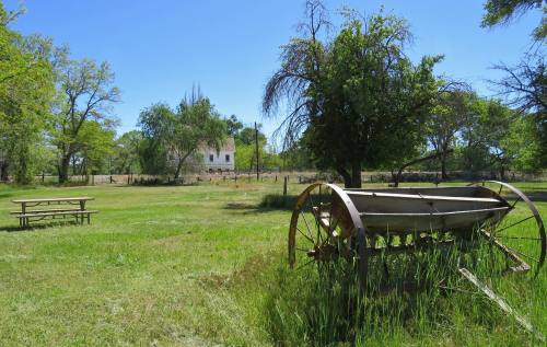 A sunny landscape featuring a vintage wagon wheel, grassy field, picnic table, and a distant house surrounded by trees.
