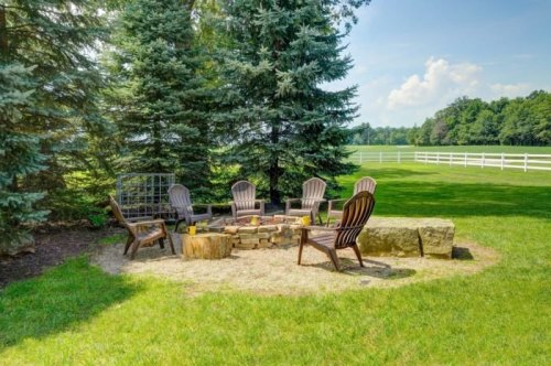 A cozy outdoor fire pit area with wooden chairs surrounded by trees and a grassy field.