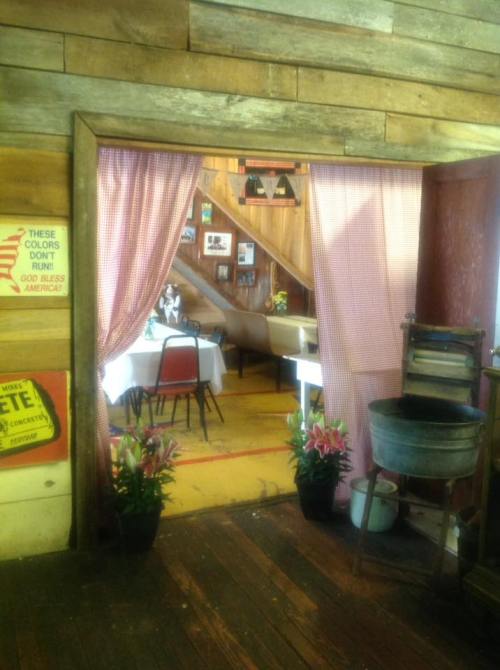 View of a rustic dining area through a doorway, featuring tables, chairs, and decorative elements in a wooden setting.