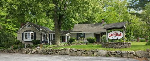 A charming gray house with a sign reading "The Daily Bean," surrounded by greenery and stone landscaping.