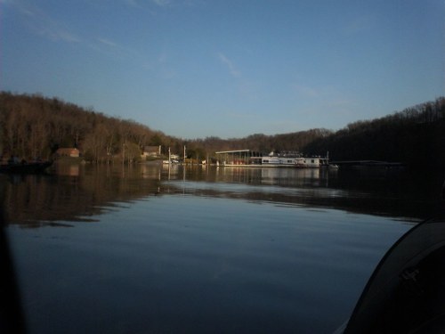 A calm lake reflecting the sky, with a marina and wooded hills in the background under a clear blue sky.