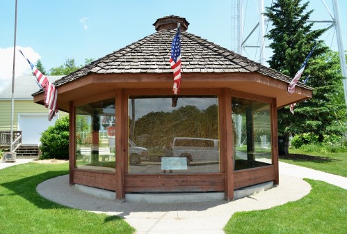 A wooden gazebo with a shingled roof, featuring glass walls and American flags, surrounded by green grass and trees.