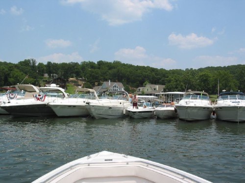 A view of several boats docked at a marina, with green hills and houses in the background under a blue sky.