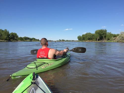 A person in a red tank top paddles a green kayak on a calm river under a clear blue sky.