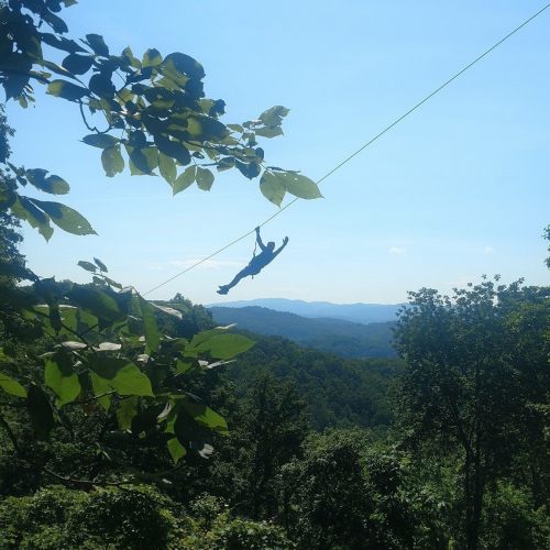 A person zip-lining over a lush green landscape with mountains in the background under a clear blue sky.
