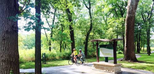Two cyclists ride along a tree-lined path in a park, with a sign for a trail visible nearby.