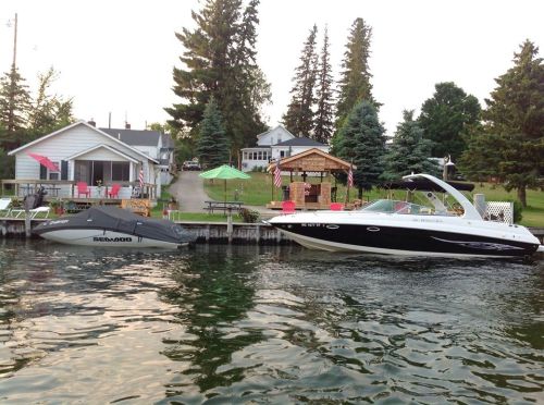 A dock with two boats, a white one and a black one, beside a lakeside house surrounded by trees.