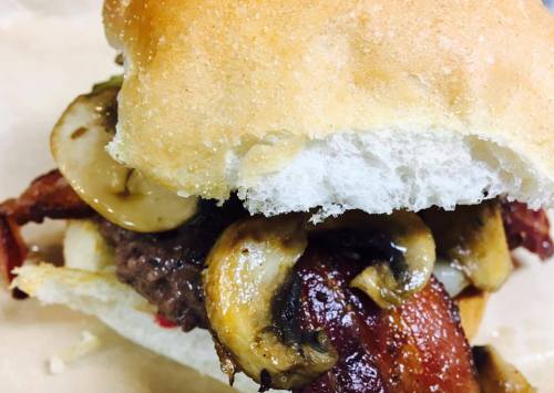 A close-up of a burger with bacon, sautéed mushrooms, and a soft bun, resting on a light-colored surface.