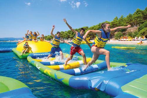 A group of kids in life jackets running and jumping on a colorful inflatable water obstacle course in a sunny beach setting.