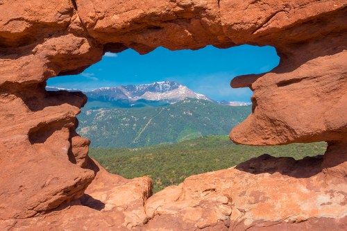 A natural rock formation frames a view of snow-capped mountains and green hills under a clear blue sky.