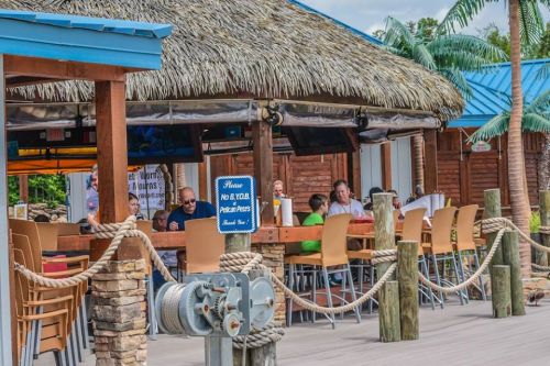 A tropical outdoor dining area with thatched roof, wooden tables, and patrons enjoying their meals. Palm trees in the background.