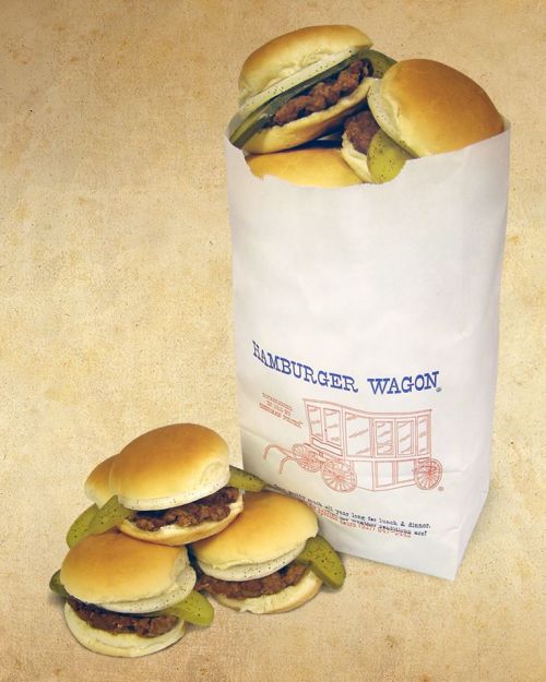 A paper bag filled with mini hamburgers and pickles, with a few burgers stacked beside it.