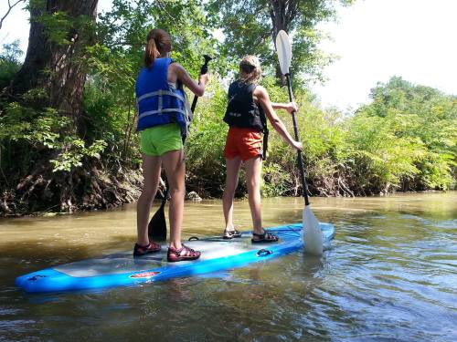 Two girls paddle on a blue stand-up paddleboard in a calm, green river surrounded by trees.