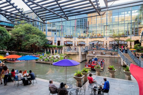 A vibrant riverside scene with colorful umbrellas, people dining, and boats on the water, surrounded by greenery and buildings.