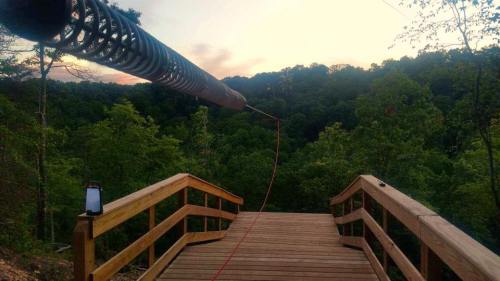 A wooden bridge leads to a scenic view of green trees and hills, with a long metal structure extending into the distance.