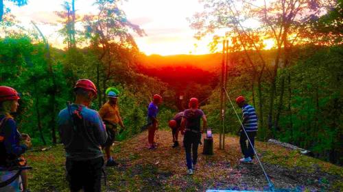A group of people in helmets stands on a hilltop at sunset, surrounded by trees and a scenic view.