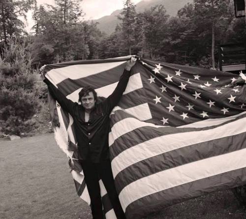 A person stands outdoors, holding a large American flag, with trees and mountains in the background.